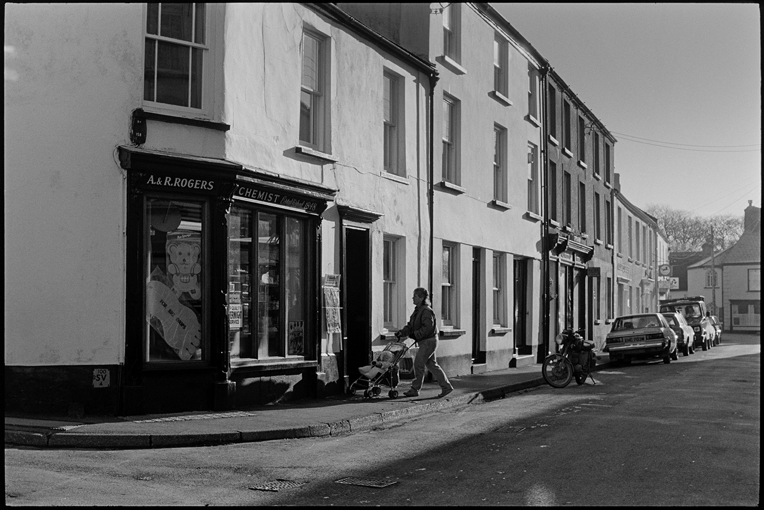 Street scene, early morning. <br /> [A woman pushing a pram past A & R Rogers Chemist in Fore Street, Chulmleigh. A motorbike and parked cars can be seen along the street in the background.]