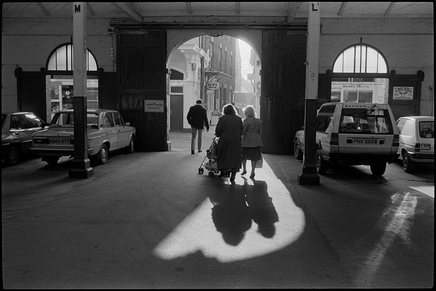 Street scenes, market building. 
[A man and two women with a pushchair leaving Barnstaple Pannier Market. Cars are parked inside the market building.]