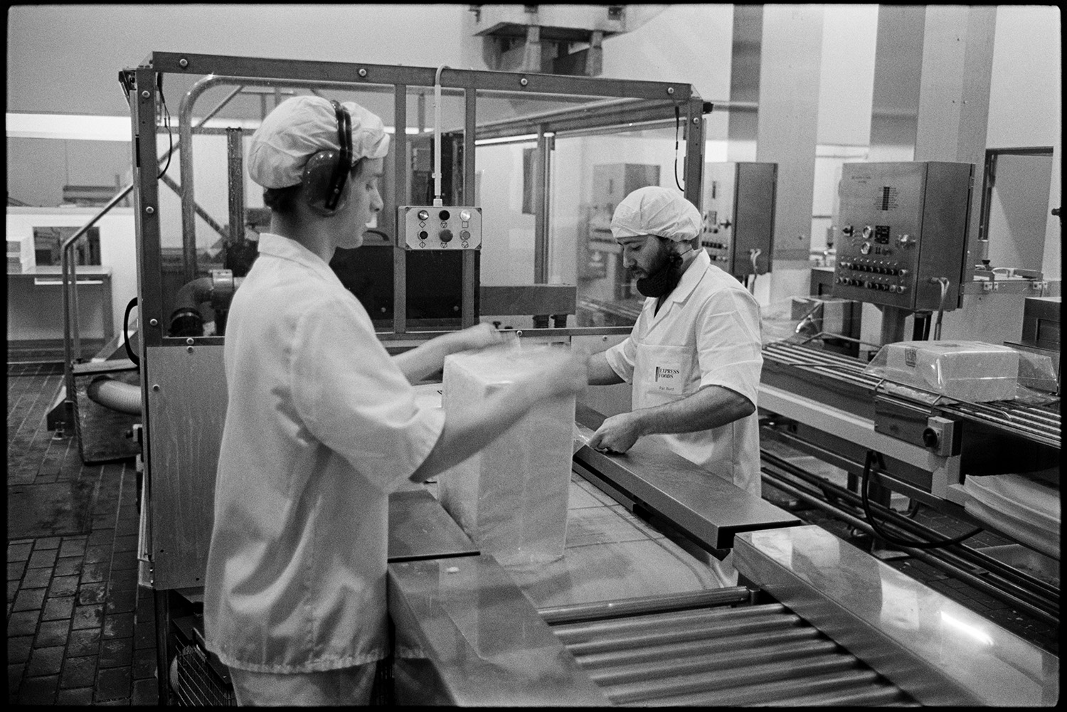 Interior of cheese factory men testing cheese and packing. 
[Two people packing cheese at the Dairy Crest North Tawton Cheese Factory. The person in the foreground is wearing ear defenders.]
