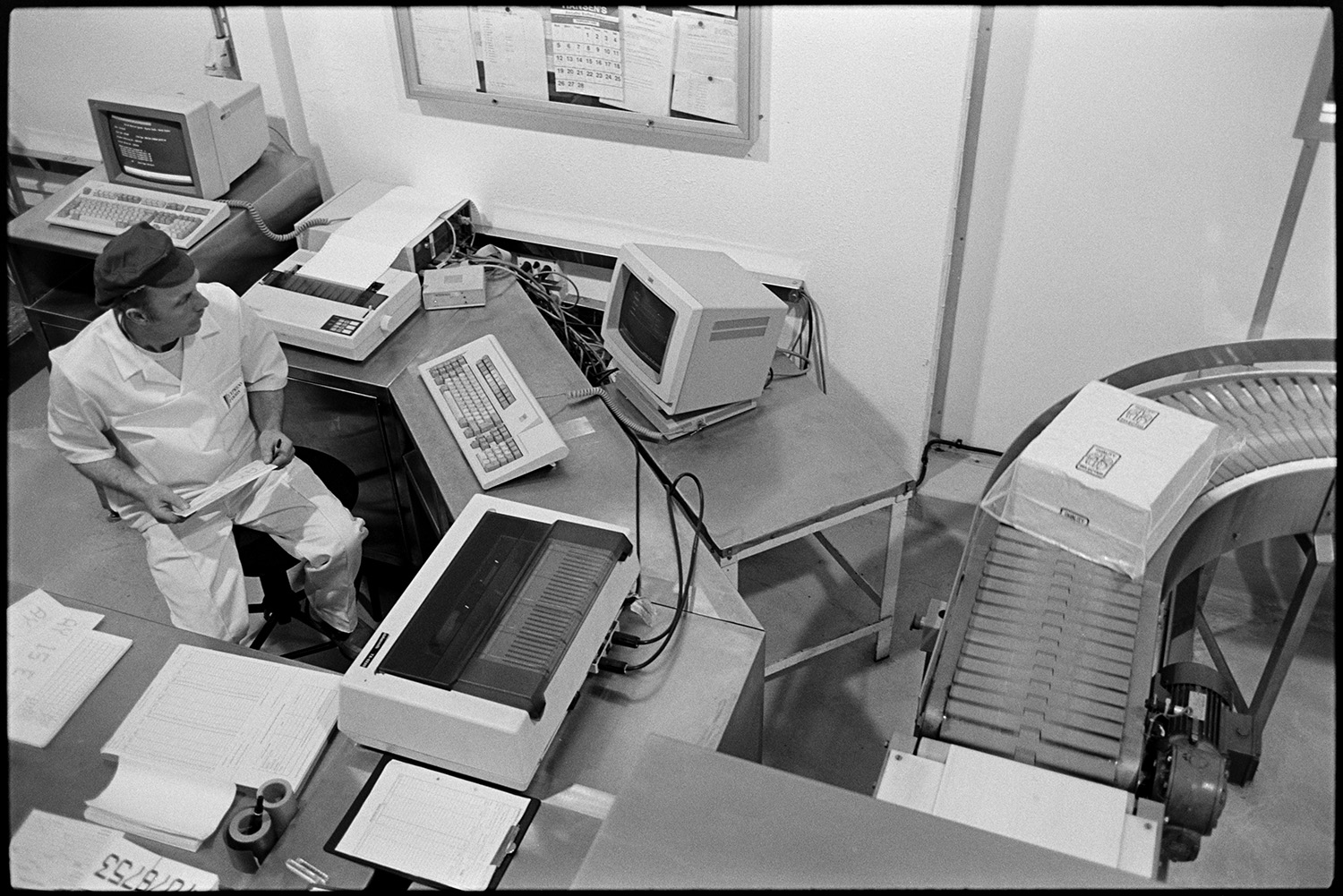Interior of cheese factory men testing cheese and packing. 
[A man sat at a desk with a computer at Dairy Crest North Tawton Cheese Factory. A conveyor belt with a box can also be seen.]