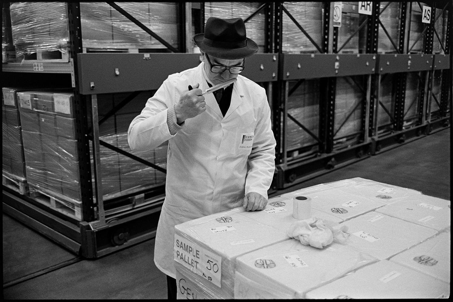 Interior of cheese factory men testing cheese and packing. 
[A man sniffing cheese from a sample pallet at the Dairy Crest North Tawton Cheese Factory. Boxes of cheese can be seen stacked in the background.]