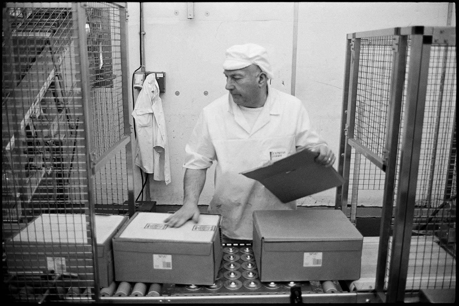 Interior of cheese factory men testing cheese and packing. 
[A man checking boxes of cheese on a conveyor belt at the Dairy Crest North Tawton Cheese Factory.]