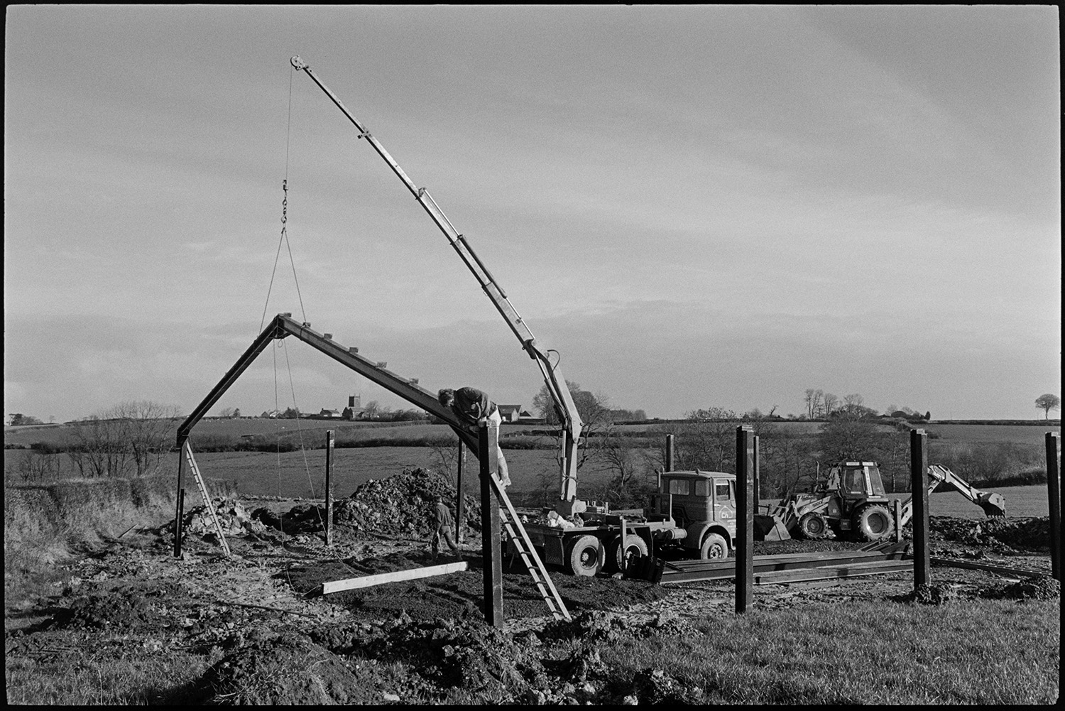 Crane erecting new barn in field. 
[A crane putting up the metal frame of a barn in a field at Ashreigney. A person is up a ladder fixing the frame together. A digger is in the background and a church tower and other buildings can be seen on the horizon.]