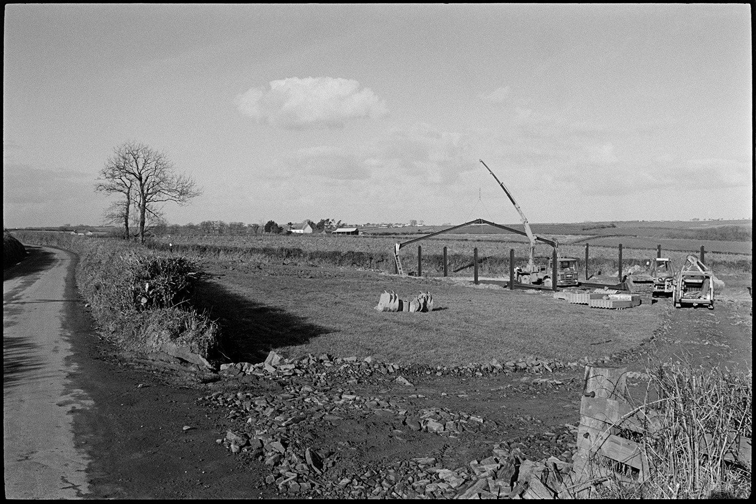 Crane erecting new barn in field. 
[A crane putting up the metal frame of a barn in a muddy field in Ashreigney.  Buckets for a digger can be seen in the foreground and farm buildings are visible in the background.]