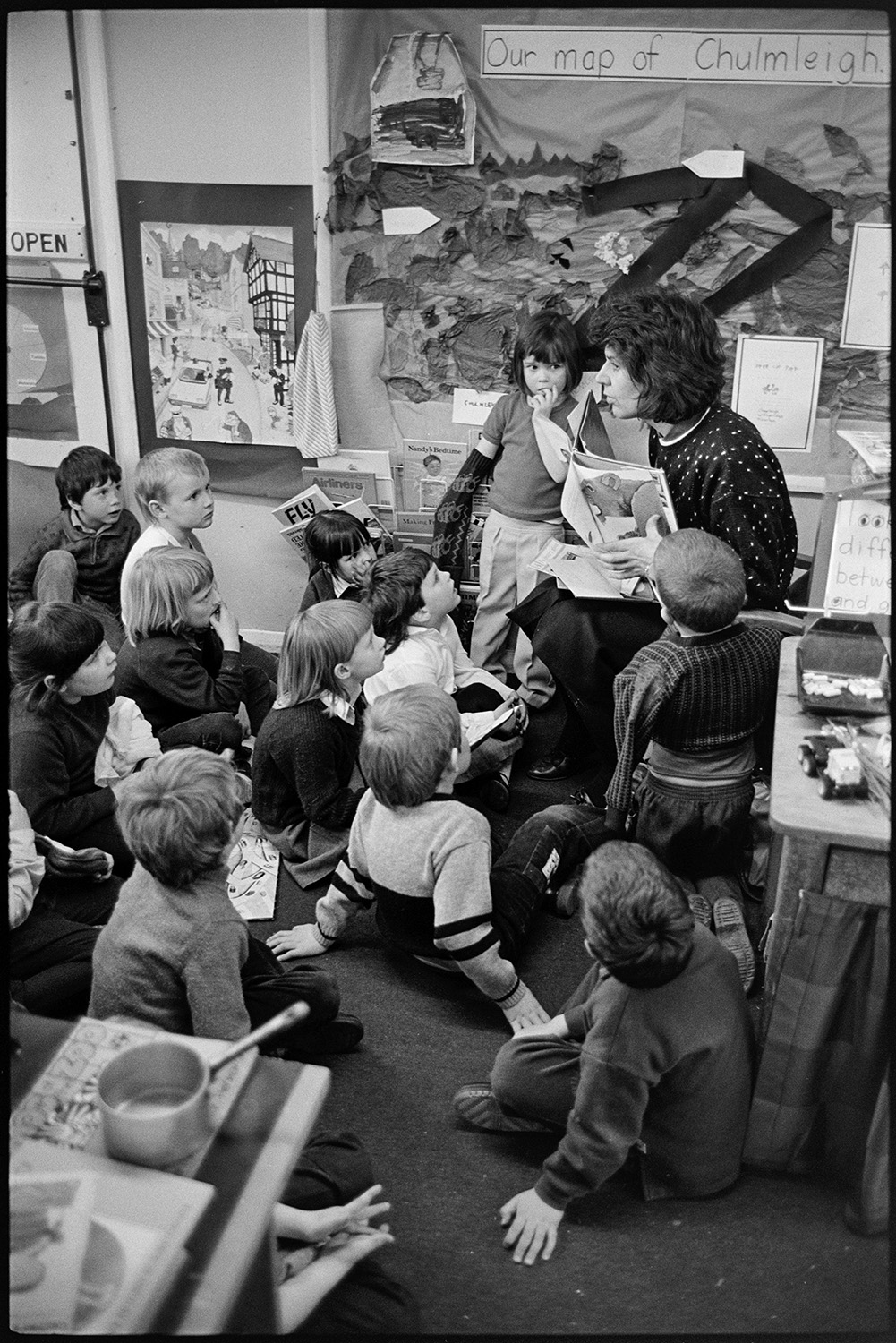 Children in primary school prefab classroom, before move to new school. 
[Mrs Bassett, a supply teacher, reading a story to children in a porta cabin classroom at Chulmleigh Primary School. Children's work is displayed on the classroom wall.]