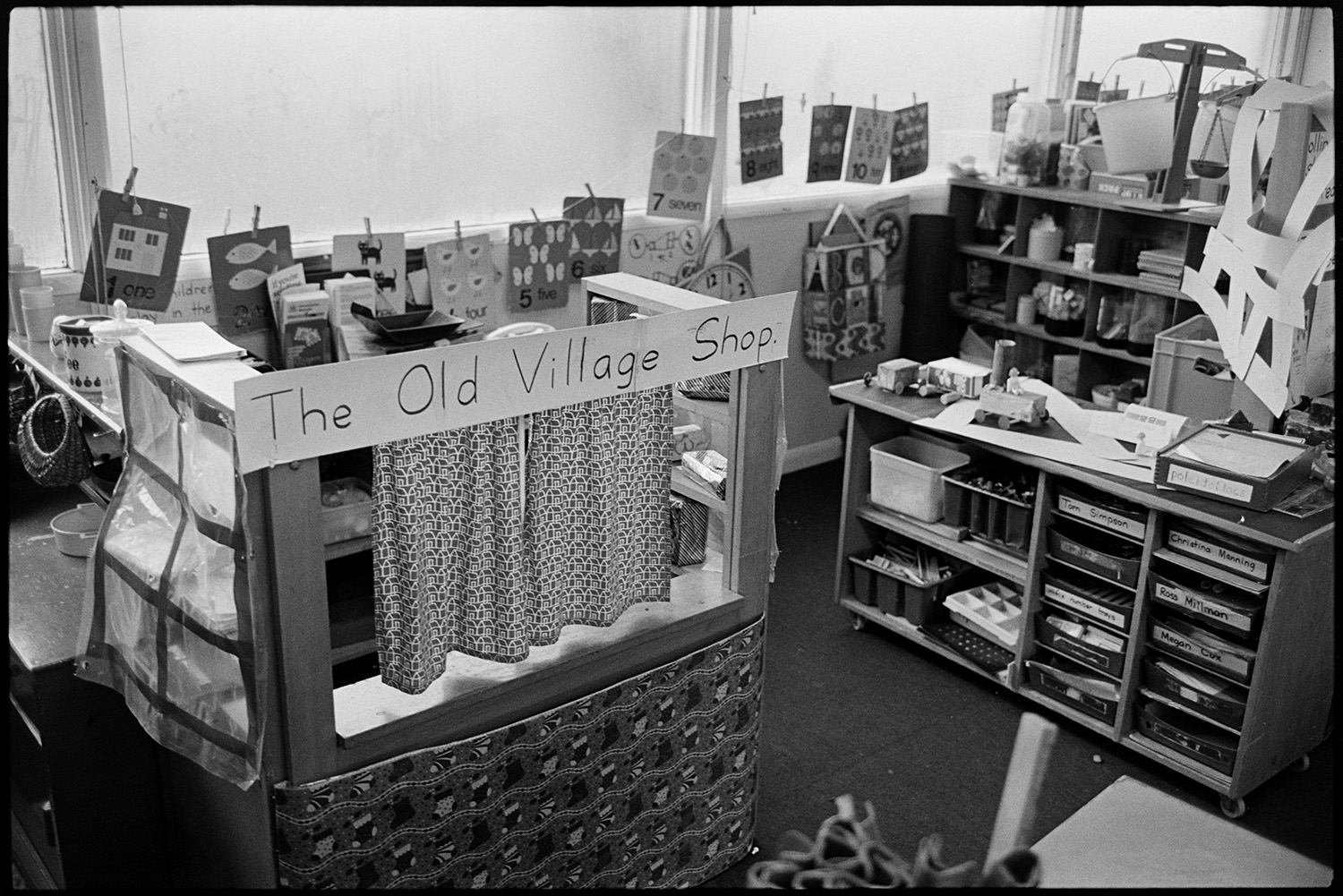 Children in primary school prefab classroom, before move to new school. 
[A classroom in a porta cabin at Chulmleigh Primary School. A small playhouse with a sign reading 'The Old Village Shop' is visible, along with a display of children's artwork and pupils' book trays.]