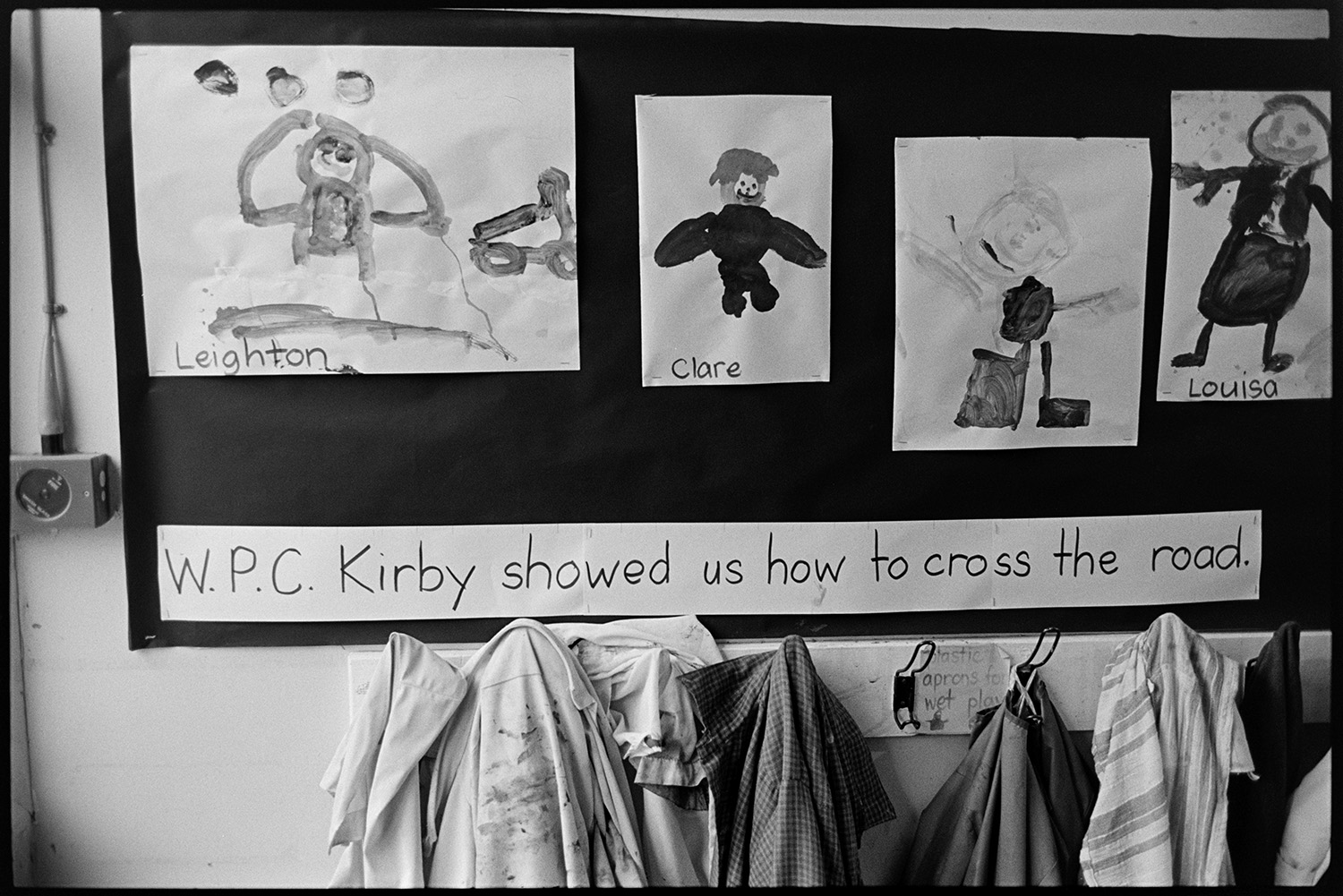 Children in primary school prefab classroom, before move to new school. 
[A display of children's artwork with the caption ' W.P.C. Kirby showed us how to cross the road' on a classroom wall at Chulmleigh Primary School. Hooks with aprons are underneath the display.]