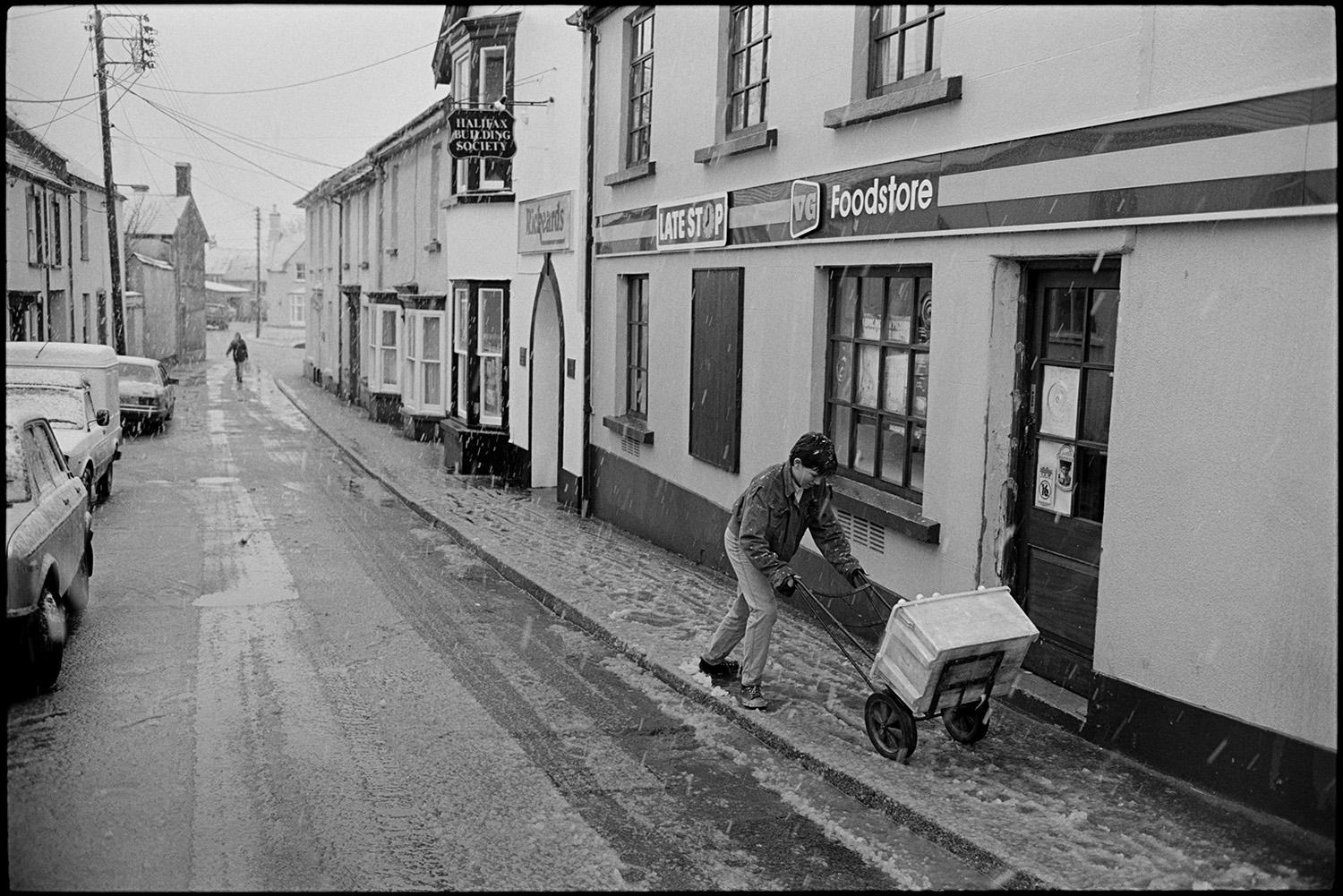 Snow, street scenes with people coming out of coffee morning. 
[A boy pushing a box on a sack truck along a snow covered pavement in Fore Street, Chulmleigh, past the Late Stop Food store shopfront. Snow is falling an settling on parked cars and the road.]