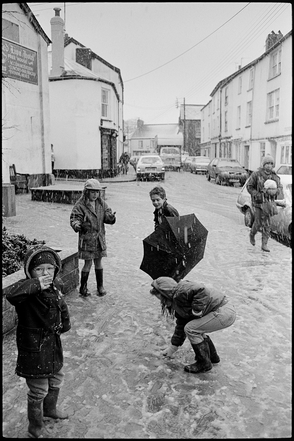 Snow, street scenes with people coming out of coffee morning. 
[Children playing in the snow in Fore Street, Chulmleigh outside the Red Lion Hotel. One of the children has an umbrella. Parked cars can be seen along the street in the background and snow is falling.]