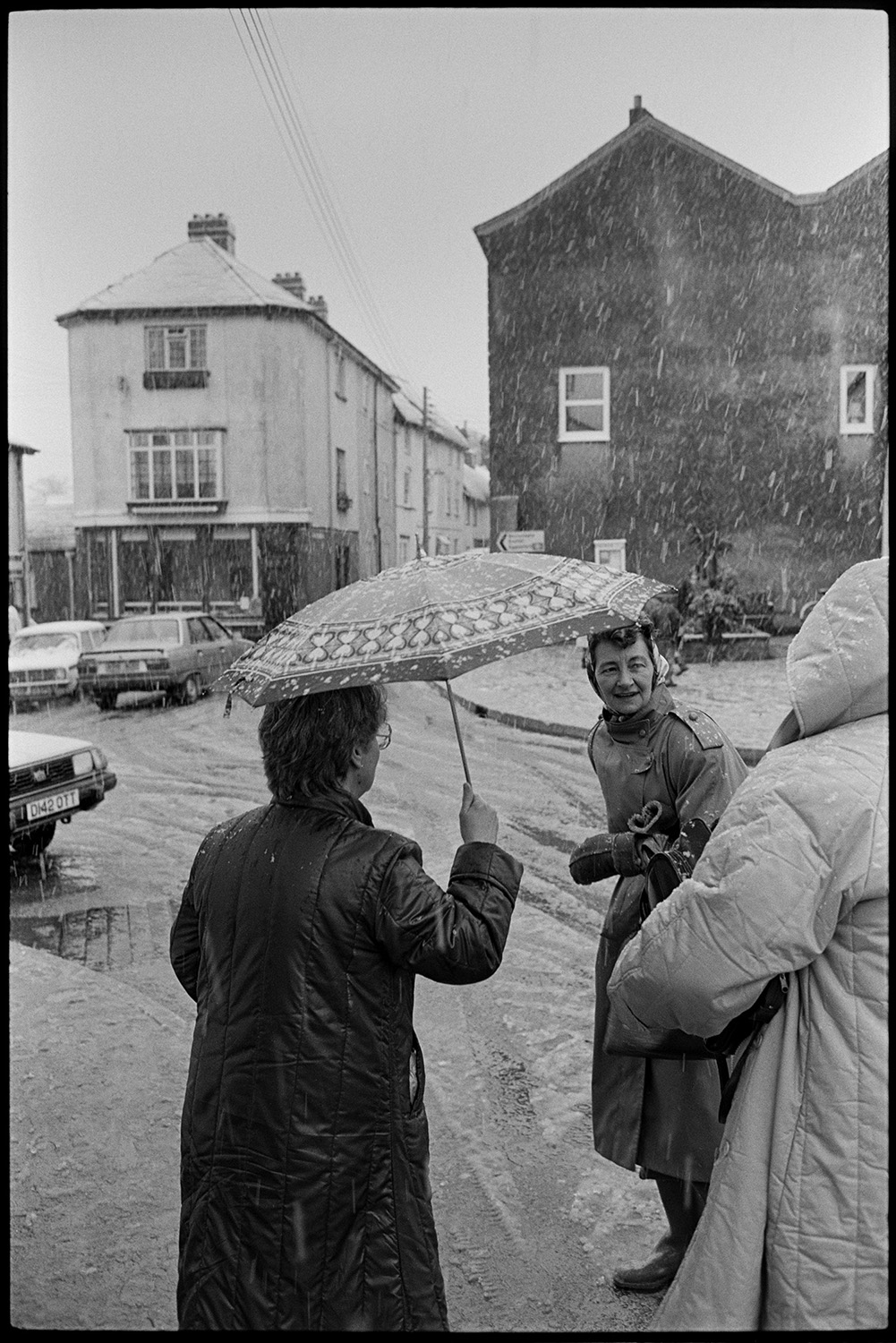 Snow, street scenes with people coming out of coffee morning. 
[Three women talking under an umbrella in Fore Street, Chulmleigh. Snow is falling and settling on the road.]