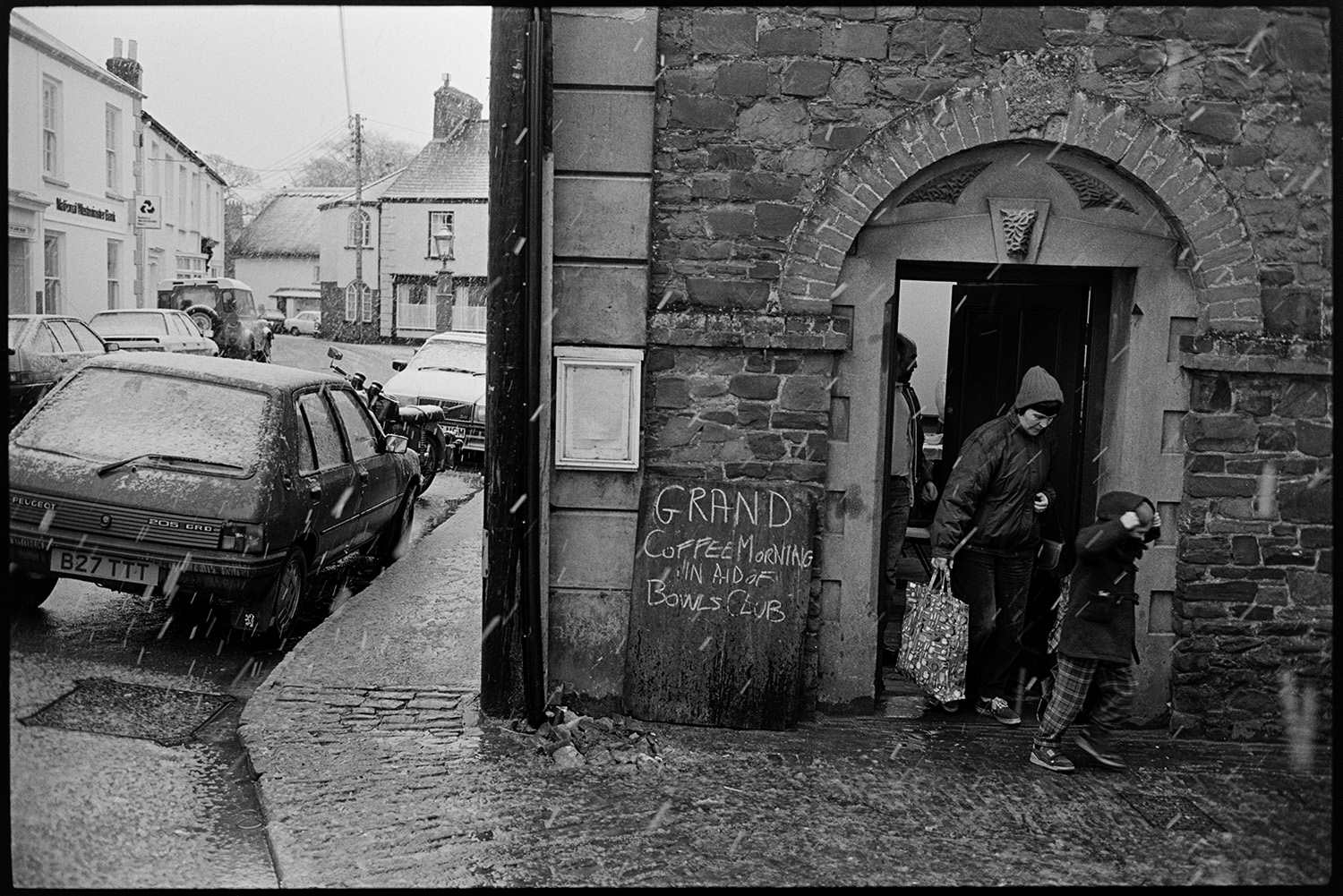 Snow, street scenes with people coming out of coffee morning. 
[A woman and child leaving a coffee morning and coming out from a building onto a cobbled street in Chulmleigh. It is snowing and the snow can be seen settling on parked cars in the adjacent street. The coffee morning was held to raise money for the Bowls Club.]