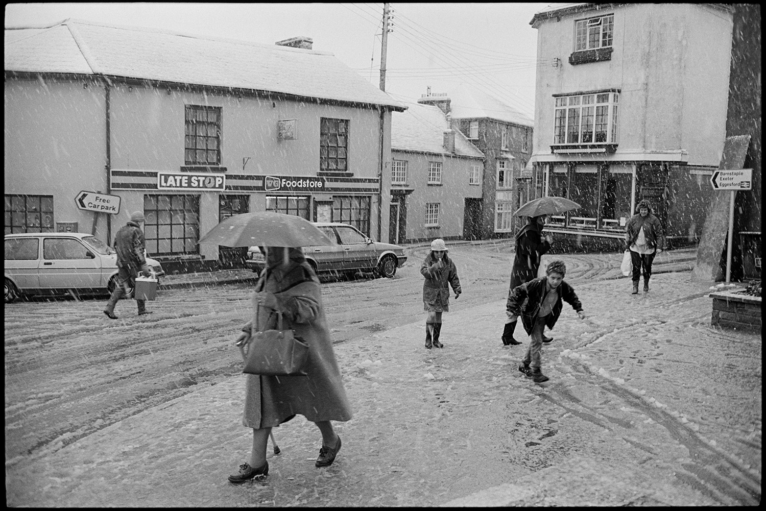 Snow, street scenes with people coming out of coffee morning. 
[People, including children, walking along Fore Street, Chulmleigh in a snow storm. Some people have umbrellas. The Late Stop Food store shop is visible I the background.]