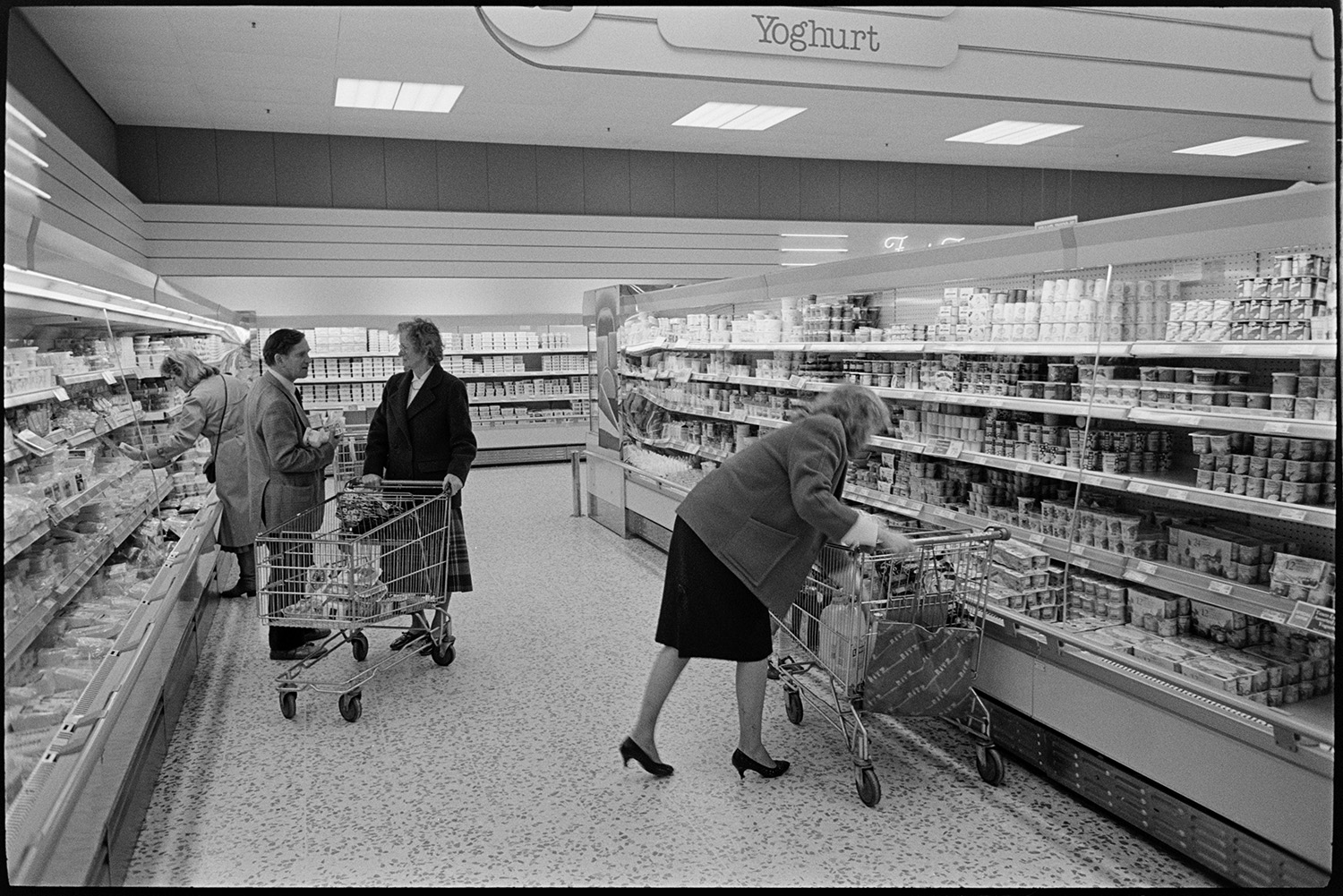 Interior of supermarket with people shopping, delivery lorry being unloaded. 
[People shopping in the Tesco supermarket in Barnstaple. Two women are selecting goods in the chilled aisle and a man and woman are talking by a trolley.]