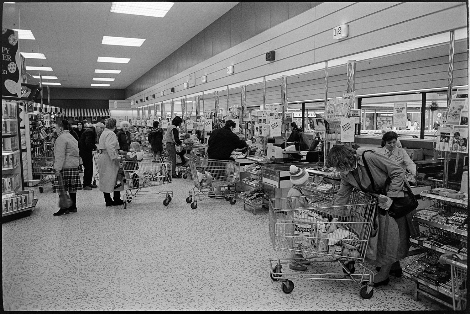 Interior of supermarket with people shopping, delivery lorry being unloaded. 
[People queuing up to buy goods at the checkouts in the Tesco supermarket in Barnstaple. A woman in the foreground is unloading a trolley with a child.]