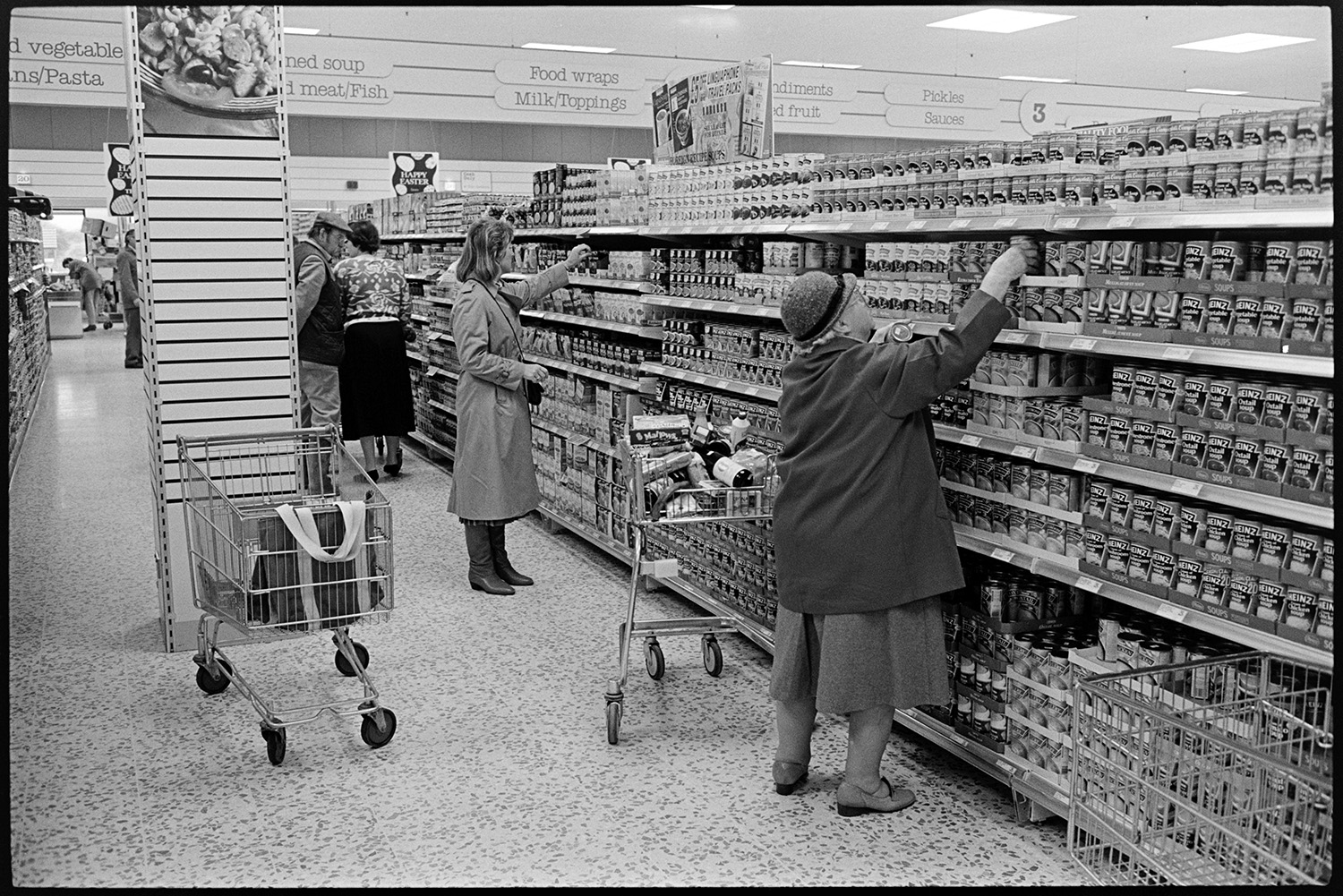 Interior of supermarket with people shopping, delivery lorry being unloaded. 
[People shopping in the Tesco supermarket in Barnstaple. Two women are selecting canned items from shelves in an aisle.]