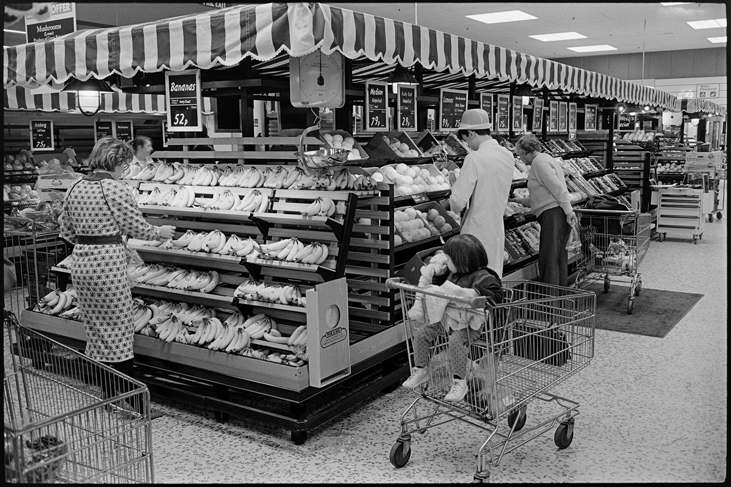 Interior of supermarket with people shopping, delivery lorry being unloaded. 
[People shopping in the Tesco supermarket in Barnstaple. A woman is selecting bananas from shelves under a striped awning while a child sits in a trolley nearby. A staff members is checking fruit further along the aisle.]