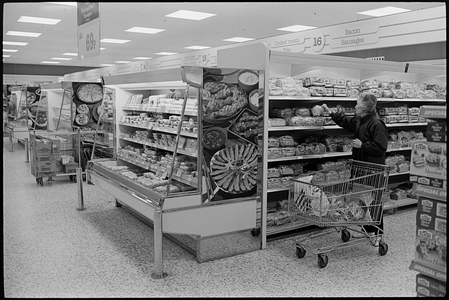 Interior of supermarket with people shopping, delivery lorry being unloaded. 
[A woman selecting a loaf of bread from a shelf on the bakery aisle in the Tesco supermarket in Barnstaple. Cabinets with chilled goods can be seen on the ends of the aisles.]