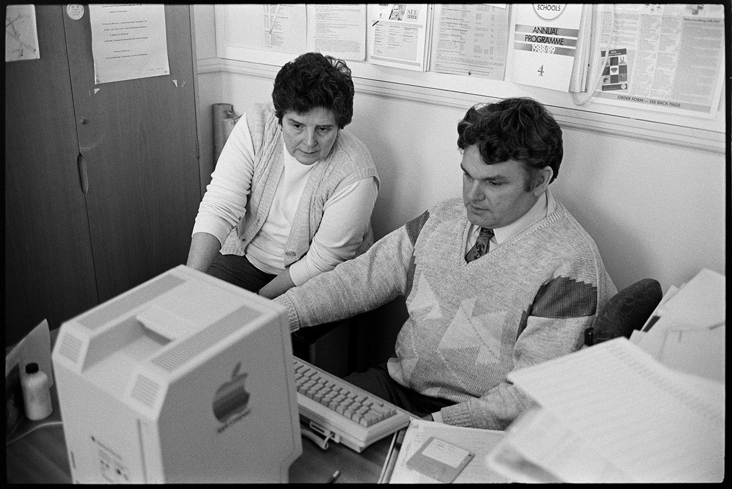 School on Red Nose Day, for Comic Relief. Headmaster sitting at desk, computer. 
[Two staff members at Chulmleigh Community College sat in an office at desk with computers. The 'Apple' brand can be seen on one of the computers.]