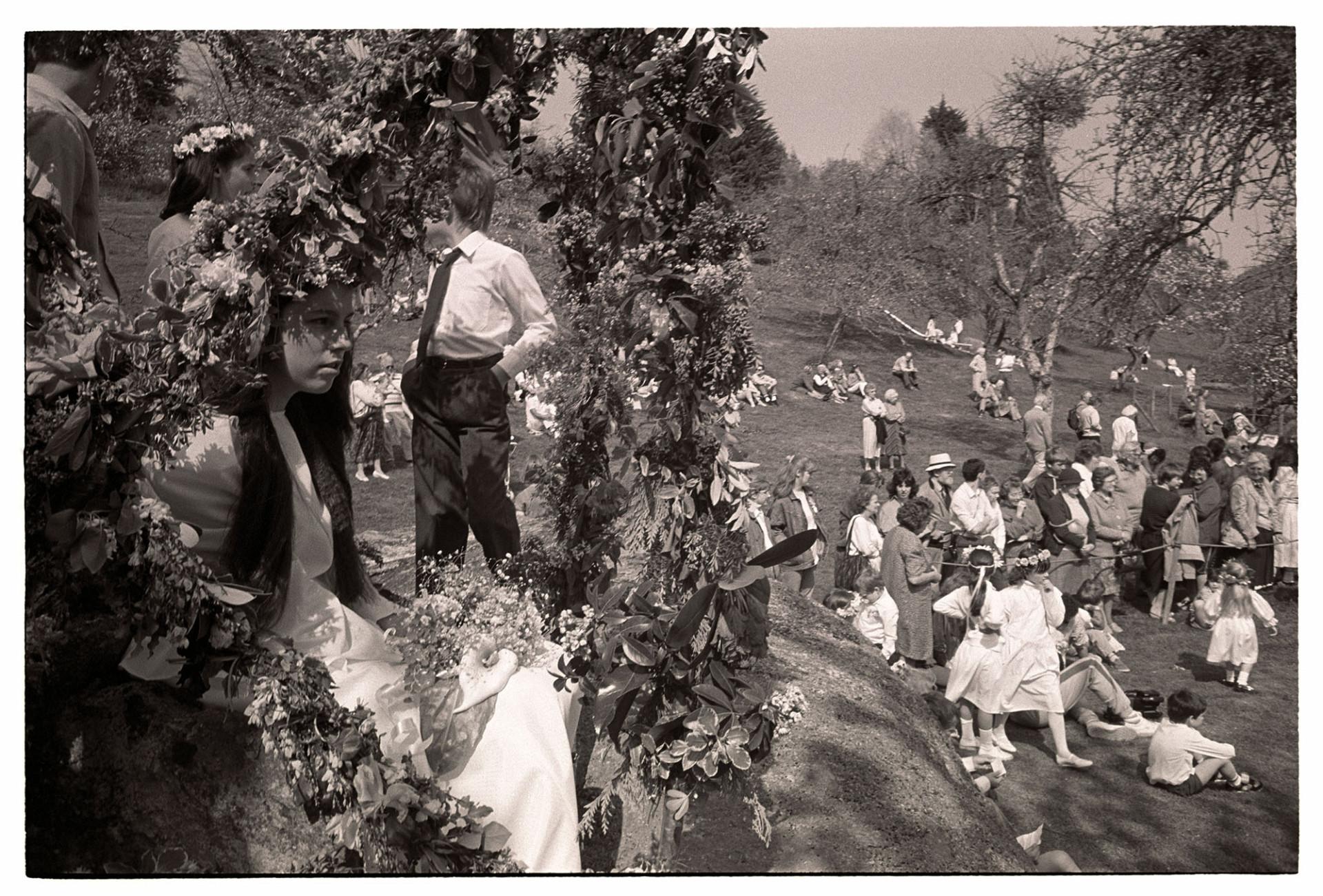 Orchards, May Queen enthroned in flower bower on rock. 
[May fair celebration in an orchard in Lustleigh. The May Queen is sat in a flower bower on a large rock. Spectators are stood and sat in the orchard. Some girls, possibly May Queen attendants, are wearing floral headbands.]