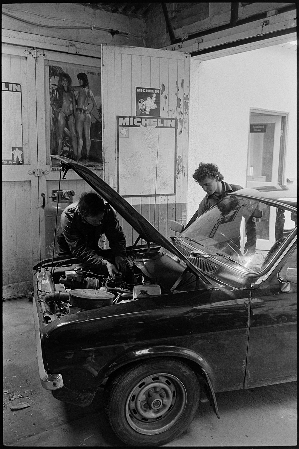 Interior of garage with car being serviced.
[A car being serviced in the workshop at the Taw Valley Garage near Eggesford by Martin Perron, who is looking under the bonnet, and another man.]