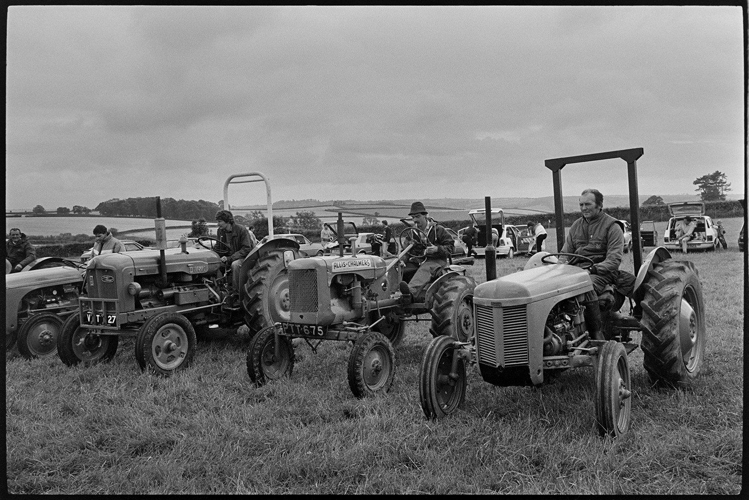 Village Club Day, sports, teas clay pigeon shoot, committee in discussion.
[A row of vintage tractors with their drivers lined up during a Village Club Day at Chawleigh. There are visitors with their cars, and fields in the background.]