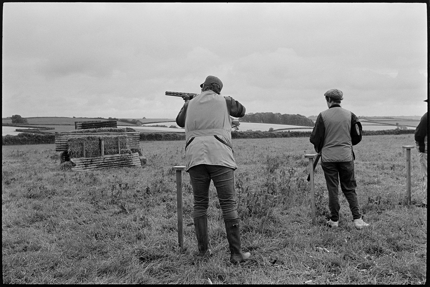 Village Club Day, sports, teas, clay pigeon shoot, committee in discussion.
[A clay pigeon shoot in a field at the Village Club Day in Chawleigh. One man is taking aim with a gun, and another is standing alongside at the next post. There are fields and woodland in the background.]