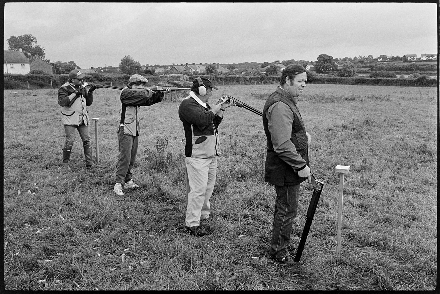Village Club Day, sports, teas, clay pigeon shoot, committee in discussion.
[A clay pigeon shoot in a field at the Village Club Day in Chawleigh. Three men are taking aim with their guns while another man is standing by the post in the foreground. There are fields and village houses in the background.]