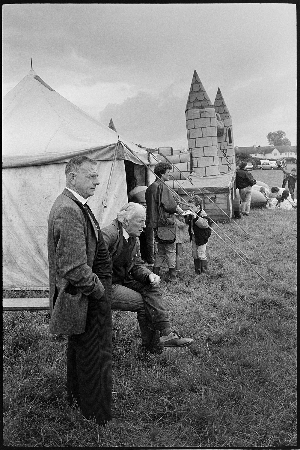 Village Club Day, sports, teas, clay pigeon shoot, committee in discussion.
[Visitors standing outside a tent during a Village Club Day at Chawleigh. There is a bouncy castle next to the tent, with some houses in the background under a stormy sky.]