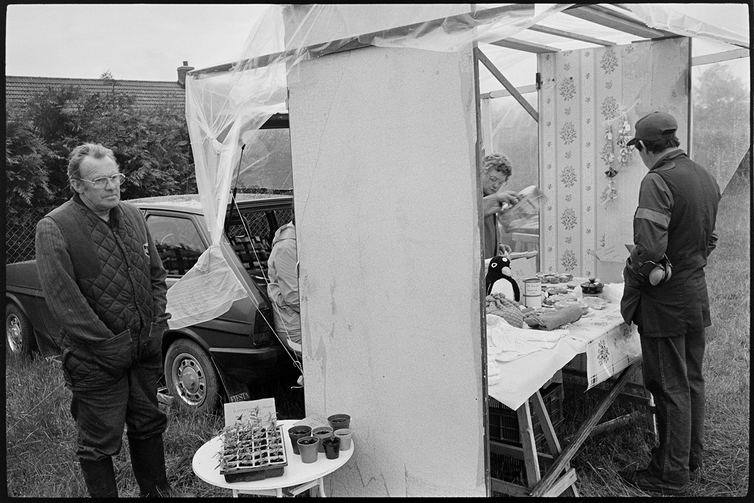 Village Club Day, sports, teas, clay pigeon shoot, committee in discussion.
[A stall at the Village Club Day at Chawleigh. A man is stood behind a small table displaying plants, next to a brick a brac stall. Another man is looking at the goods on the stall. A polythene sheet covers the stall and the car behind it.]