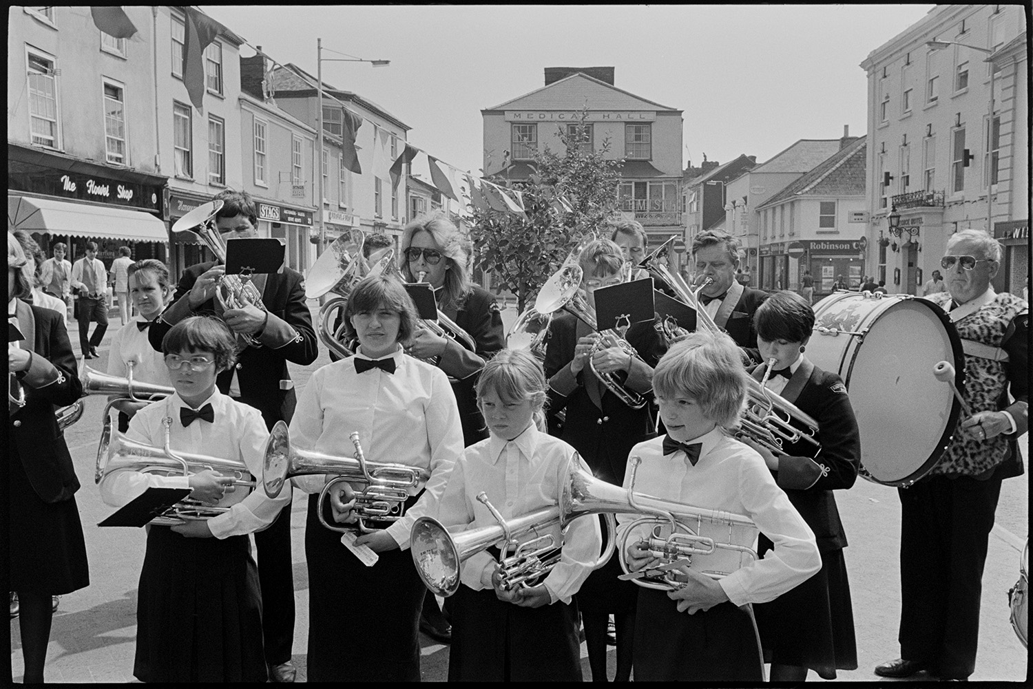 Band playing in village.
[The South Molton Town Band playing in the square in South Molton. They are in uniform and playing various brass instruments along with a bass drum. Bunting is attached to a small tree behind the band, and a number of shops and town houses are in the background.]