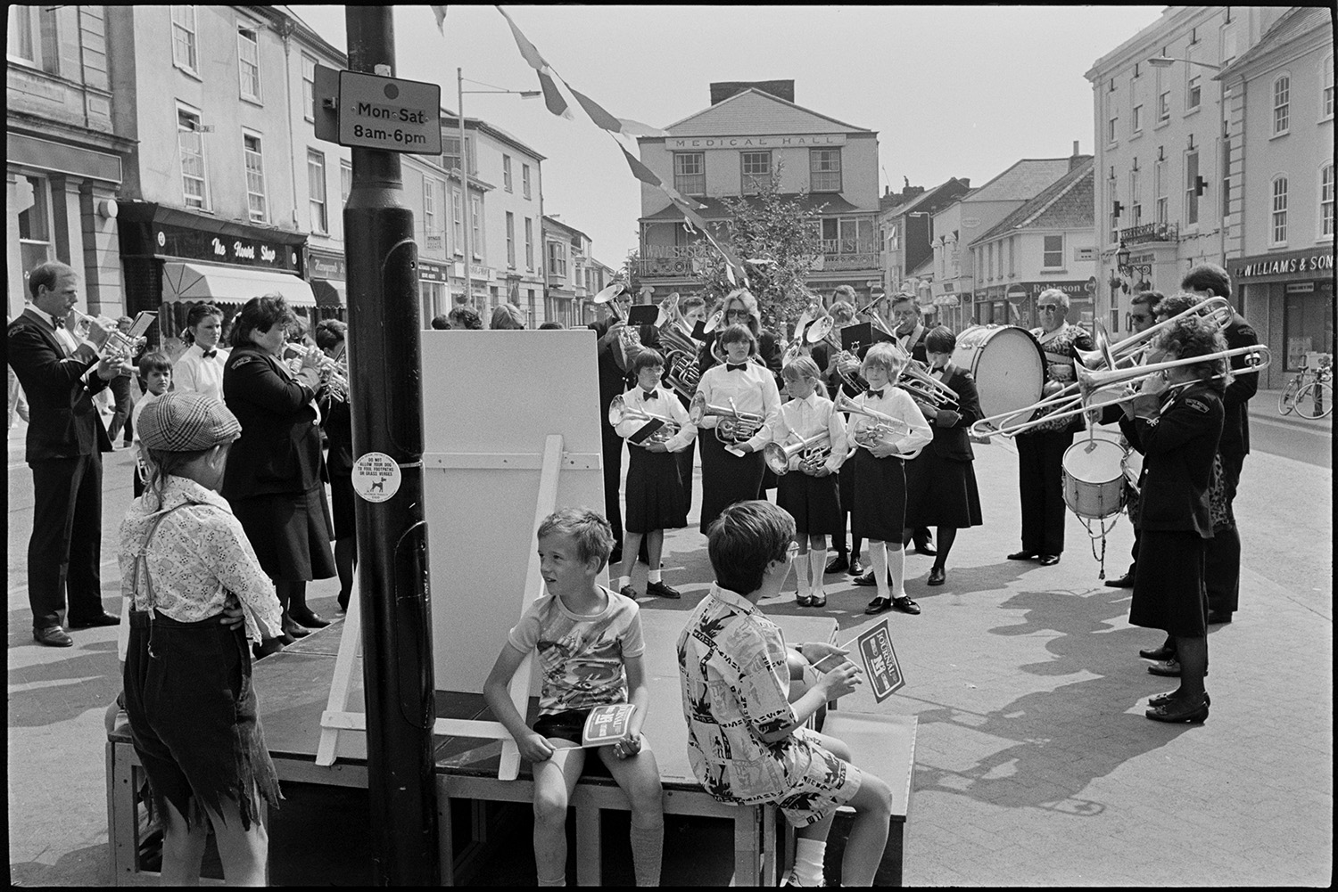 Band playing in village.
[The South Molton Town Band playing in the square in South Molton. They are in uniform and playing various brass instruments and drums. Three children are in front of them, two sitting down beside a notice board and holding small flags, and another is in fancy dress. Bunting is attached to a small tree behind the band, and a number of shops and town houses are in the background.]