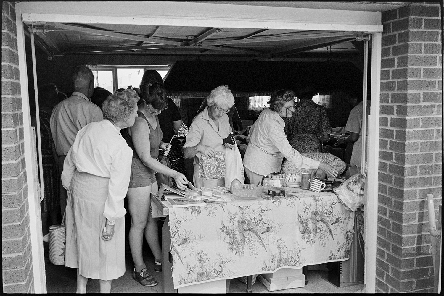 Church fete in garden, stalls, food and cakes.
[Visitors at a stall located in an open garage during a Church fete at Cricket Close, Chulmleigh. Four women are looking through goods on a bric-a-brac stall.]
