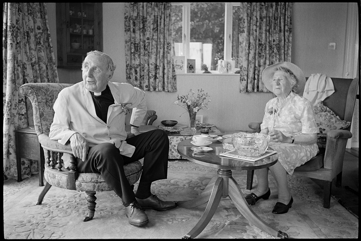 Church fete in garden, stalls, cakes, vicar making speech, thatched pavilion.
[The Reverend Richards of Chulmleigh and a woman having tea at the church fete, in a sitting room in a house at Cricket Close, Chulmleigh. There are two armchairs, a small round table, and floral curtains in the room.]
