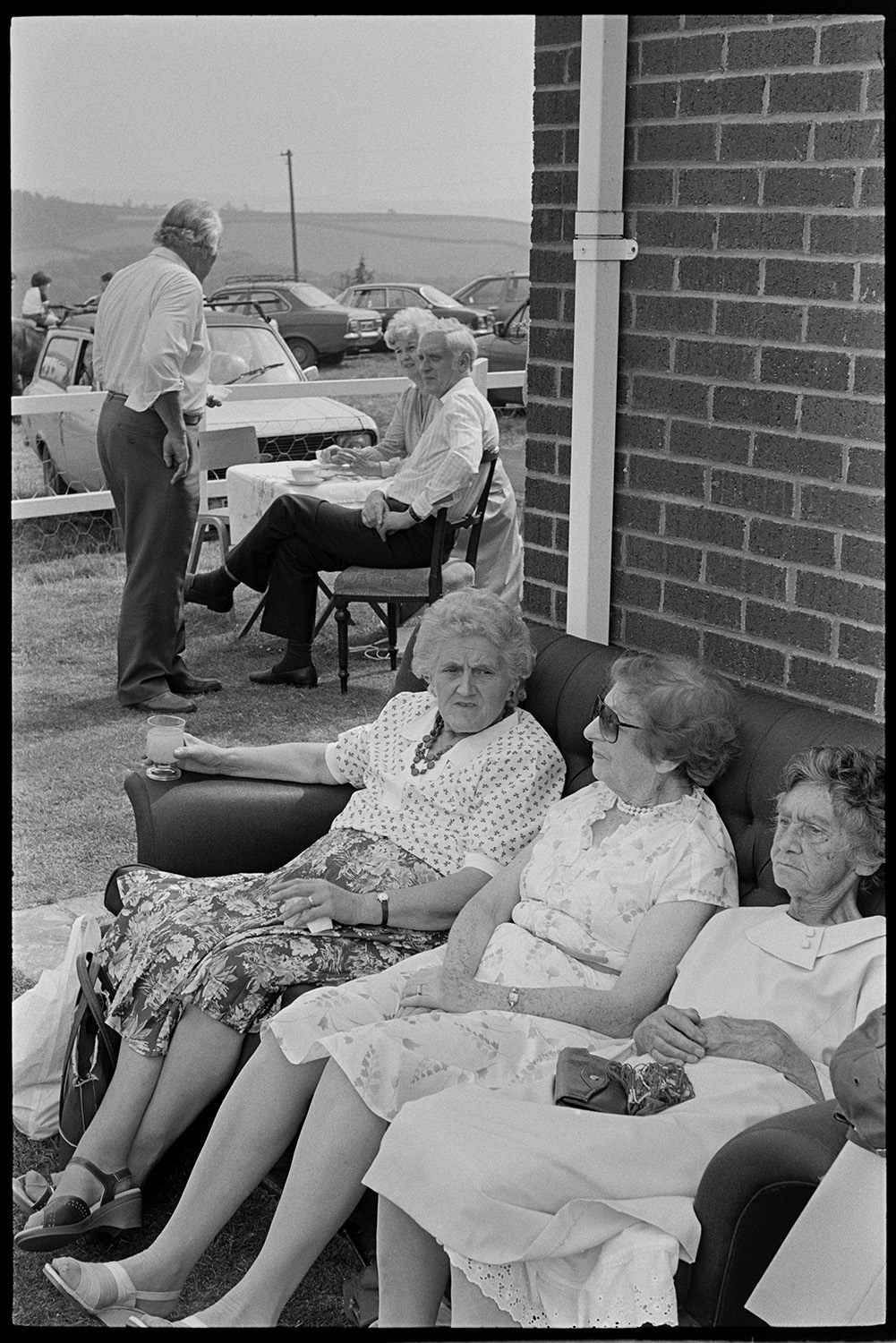 Church fete in garden, stalls, cakes, vicar making speech, thatched pavilion.
[Three elderly women sitting on an outside sofa at a church fete at Cricket Close, Chulmleigh. There are other visitors having tea at a small table, behind them, with parked cars and fields in the background.]