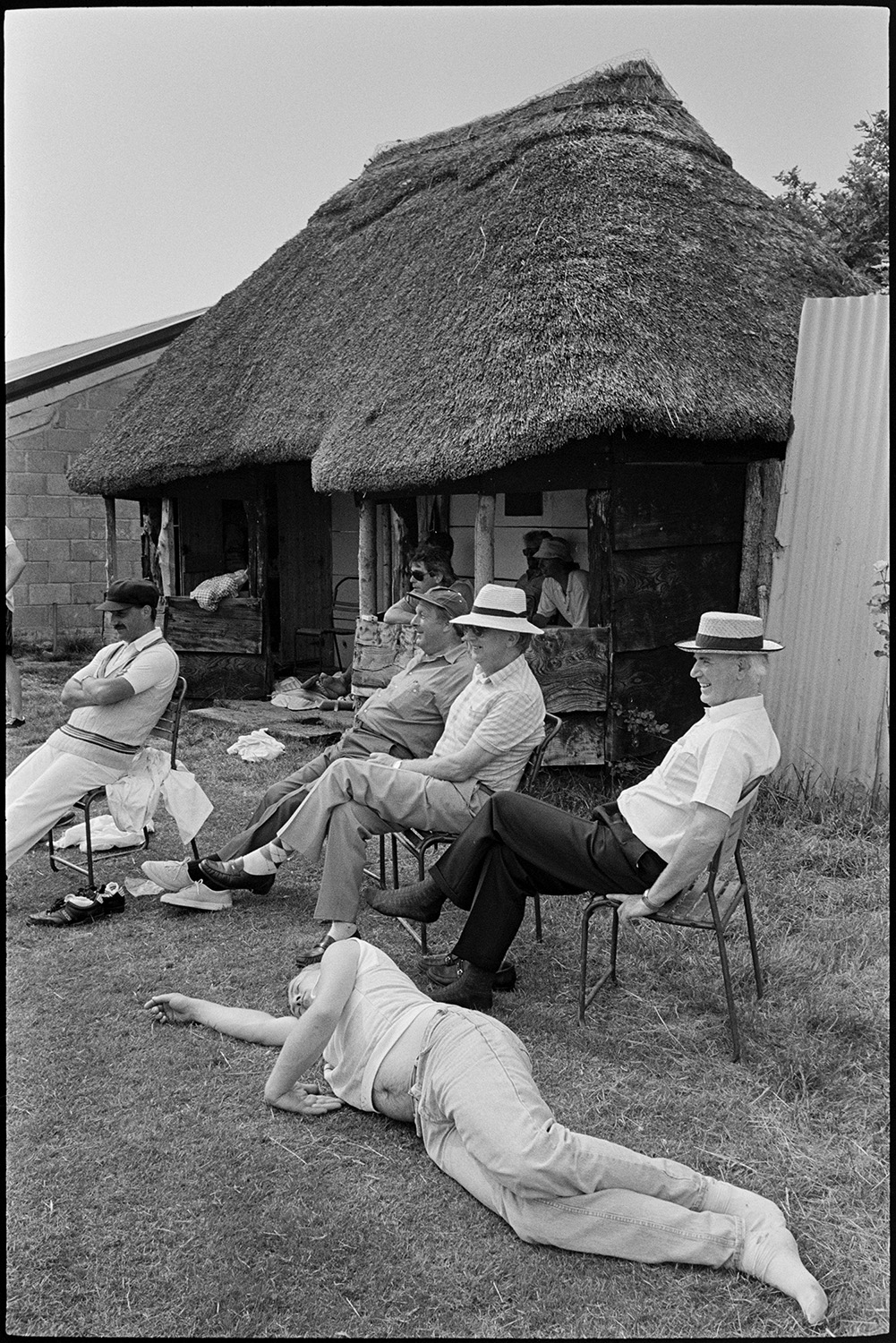 Church fete in garden, stalls, cakes, vicar making speech, thatched pavilion.
[Cricket players and spectators sitting and watching a match on the boundary at Chulmleigh cricket ground in Cricket Close, Chulmleigh, at the church fete. One man is lying on the ground asleep. Others are sitting inside the thatched and wooden framed pavilion.]