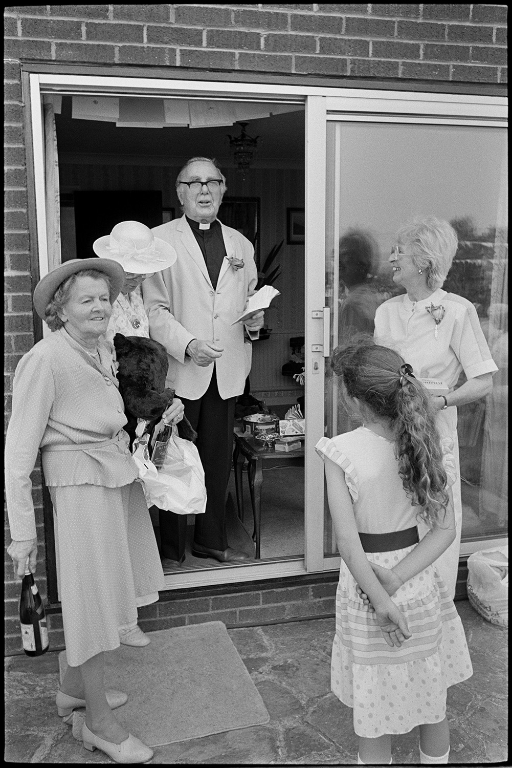 Church fete in garden, stalls, cakes, vicar making speech, thatched pavilion.
[Reverend Richards of Chulmleigh is holding a notebook at the patio door of a house during a church garden fete, at Cricket Close, Chulmleigh. There is a young girl with a ponytail, and three women holding raffle prizes, including a stuffed toy and bottles of wine.]