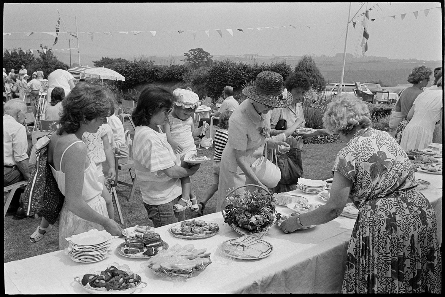 Church fete in garden, stalls, cakes, vicar making speech, thatched pavilion.
[Women and children at a refreshments table during a Church fete in a garden at Cricket Close, Chulmleigh. There are plates, cakes and sandwiches on the table. People sitting at tables, with parked cars behind a hedge, with flags and bunting in the background.]