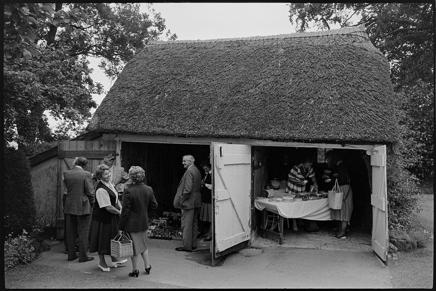 Open garden, with cake stall and people looking around garden. Thatched garage.
[Visitors at an open garden event at Highdown, Ashreigney, hosted by Moggie Dew. There is a thatched double garage with open wooden doors. One garage has potted plants on the floor, the other garage has a table selling cakes.]