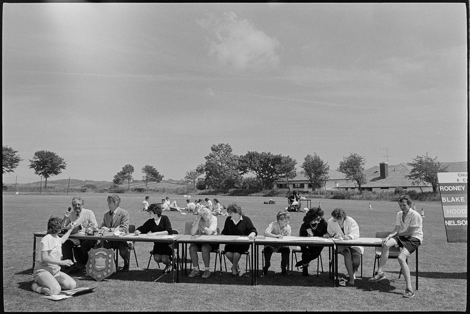School sports, pupils gathered for prize giving, crowd.
[An announcer with a microphone and scorekeepers sitting at a table during a sports day at Chulmleigh Community College. There is a scoreboard alongside the table, with a shield and trophies on display. Pupils are sitting on the grass behind the table and some of the school building is visible in the background.]