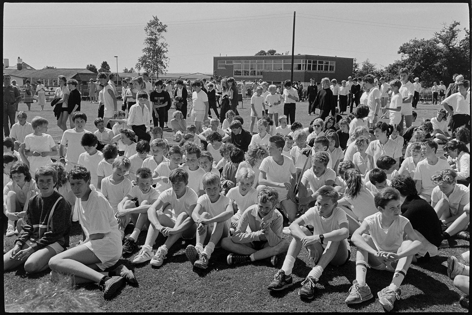 School sports, pupils gathered for prize giving, crowd.
[School pupils are gathered for a prize giving event after the school sports day at Chulmleigh Community College. Most are sitting on the grass in white t-shirts. More students and school buildings are visible in the background.]