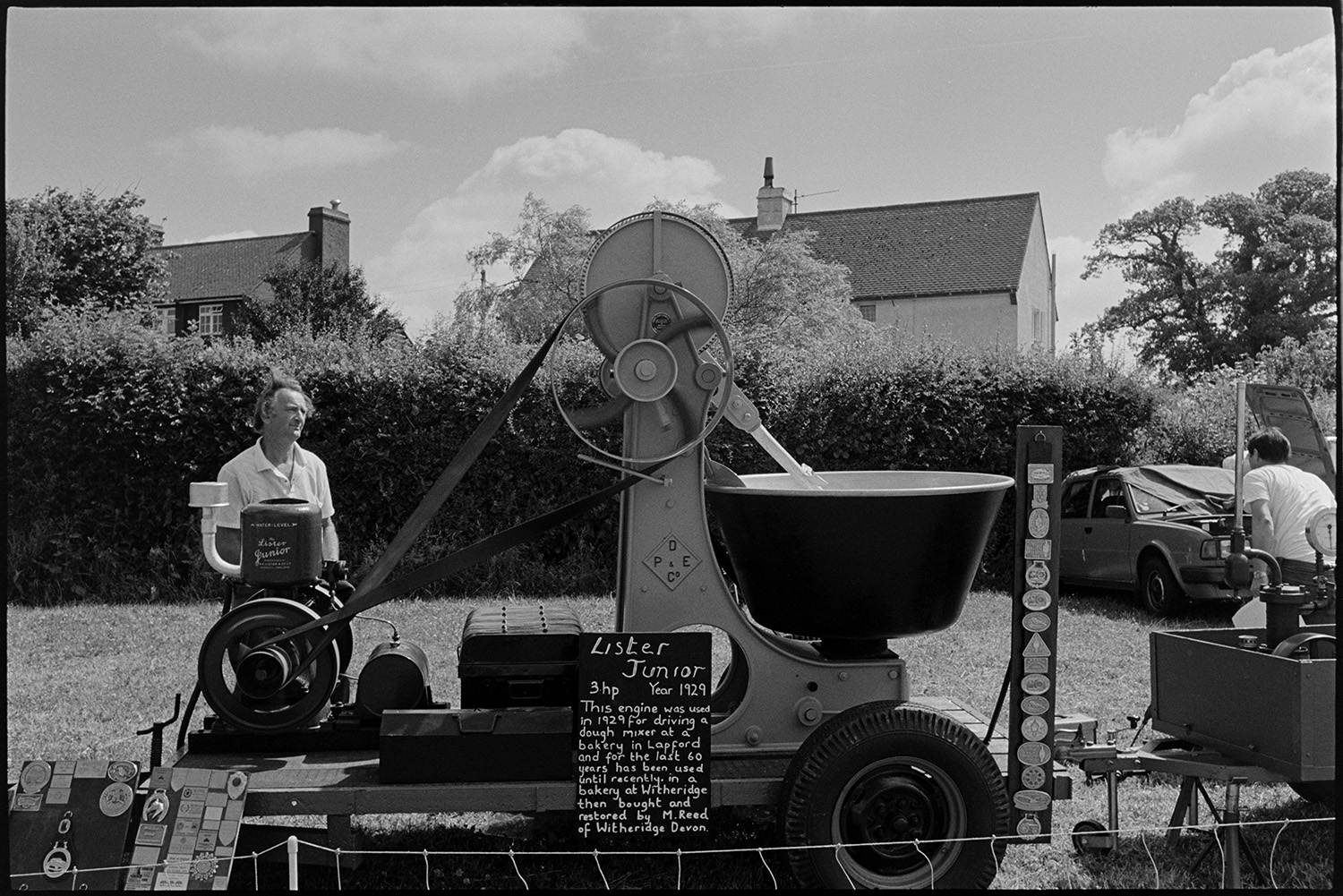 Country fair, vintage cars, sheep being rounded up for show.
[A Lister Junior diesel engine on display in a field at Ashreigney Country Fair. Behind it is a tall hedge, trees and village houses.]