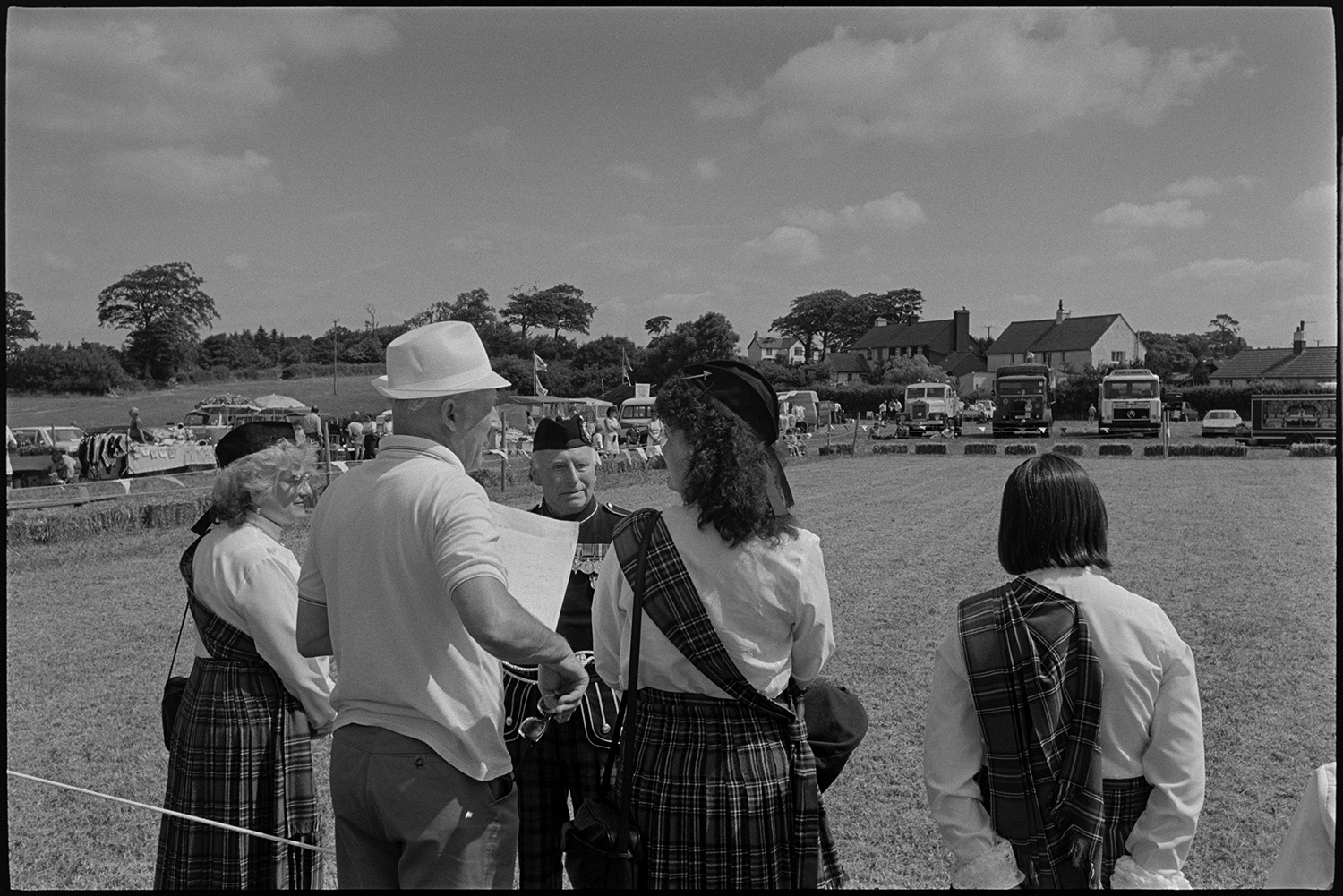 Country fair, vintage cars, sheep being rounded up for show.
[Three woman dressed in tartan, and a man in uniform at  Ashreigney Country Fair. There is a parade ring bordered by straw bales, with various stalls and vintage vehicles on display around it.]