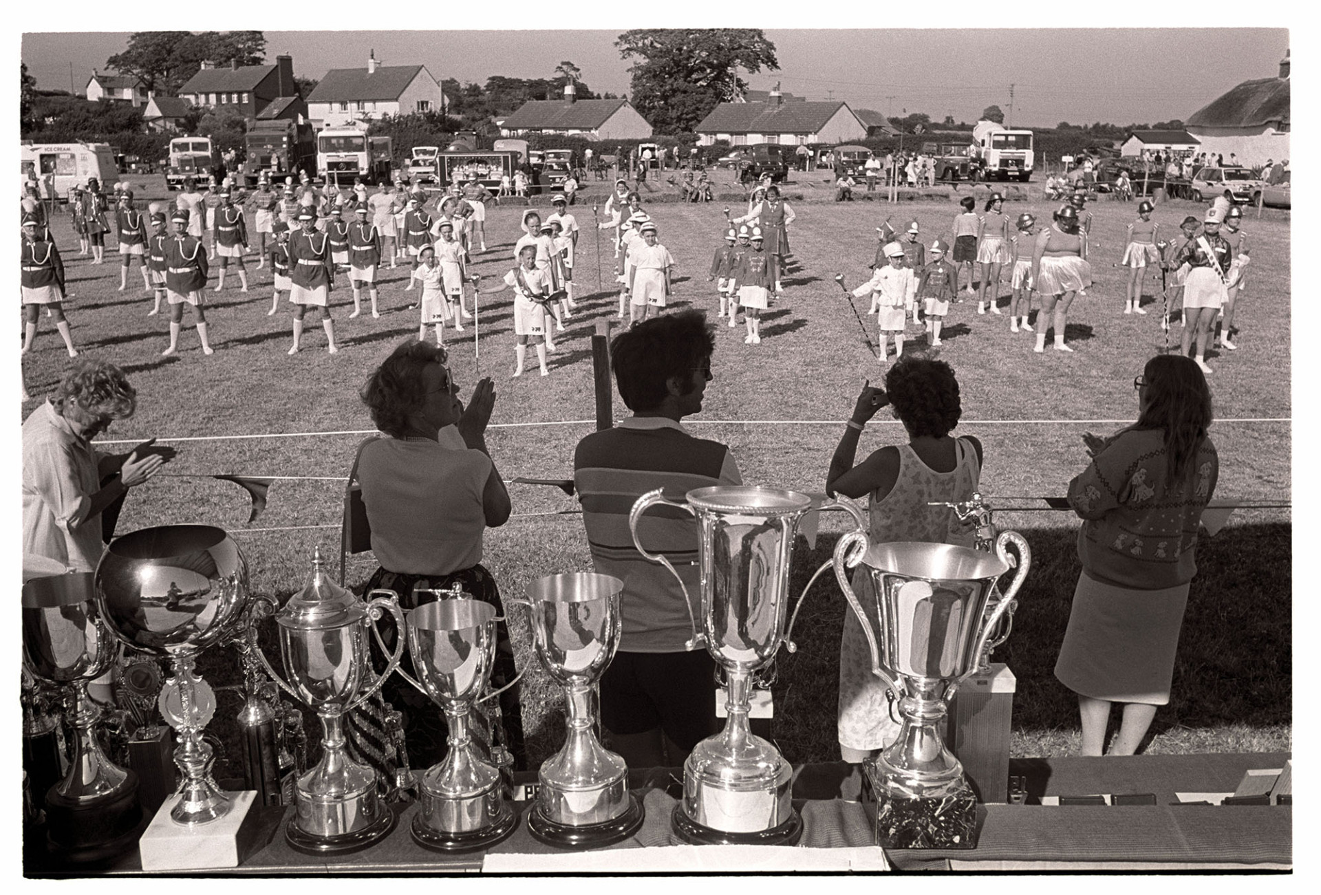 Country fair, majorettes, cups on display. 
[People watching and clapping majorettes performing at Ashreigney Country Fair. A table with trophies is in the foreground and various trucks and cars are parked in the background.]