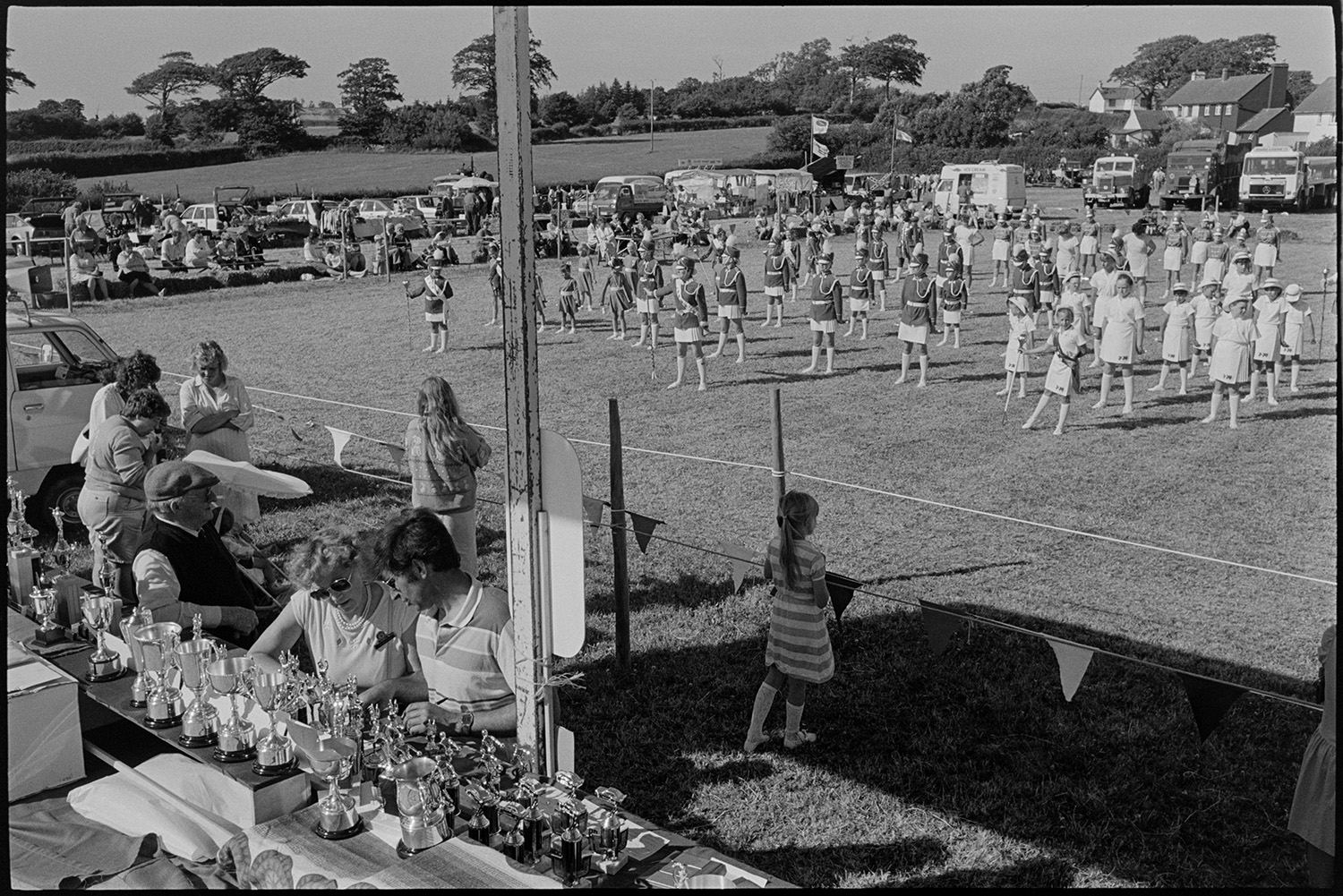 Country fair, majorettes, cups on display, stalls.
[Spectators watching groups of majorettes assembled in the showground at Ashreigney Country Fair. There is a table with trophies on display. Also in the field are spectators, vintage lorries, stalls and an ice cream van.]
