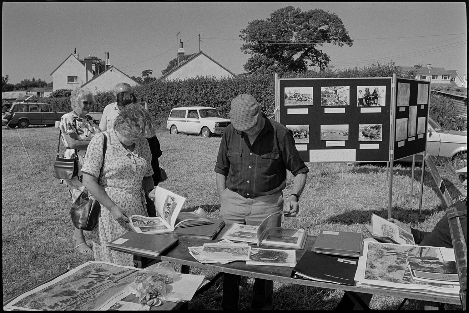 Country fair, majorettes, cups on display, stalls.
[Men and women looking through photographs from the Beaford Archive displayed on an outside table at Ashreigney Country Fair. There are display boards with more photographs, parked cars, and some village houses in the background.]
