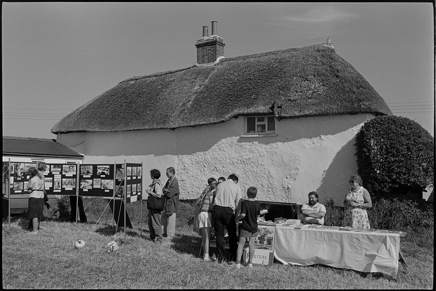 Country fair, majorettes, cups on display, stalls.
[Visitors looking at display boards showing a range of photographs from the Beaford Archive, and a stall selling posters, at Ashreigney Country Fair. They are in front of a whitewashed and thatched cottage.]
