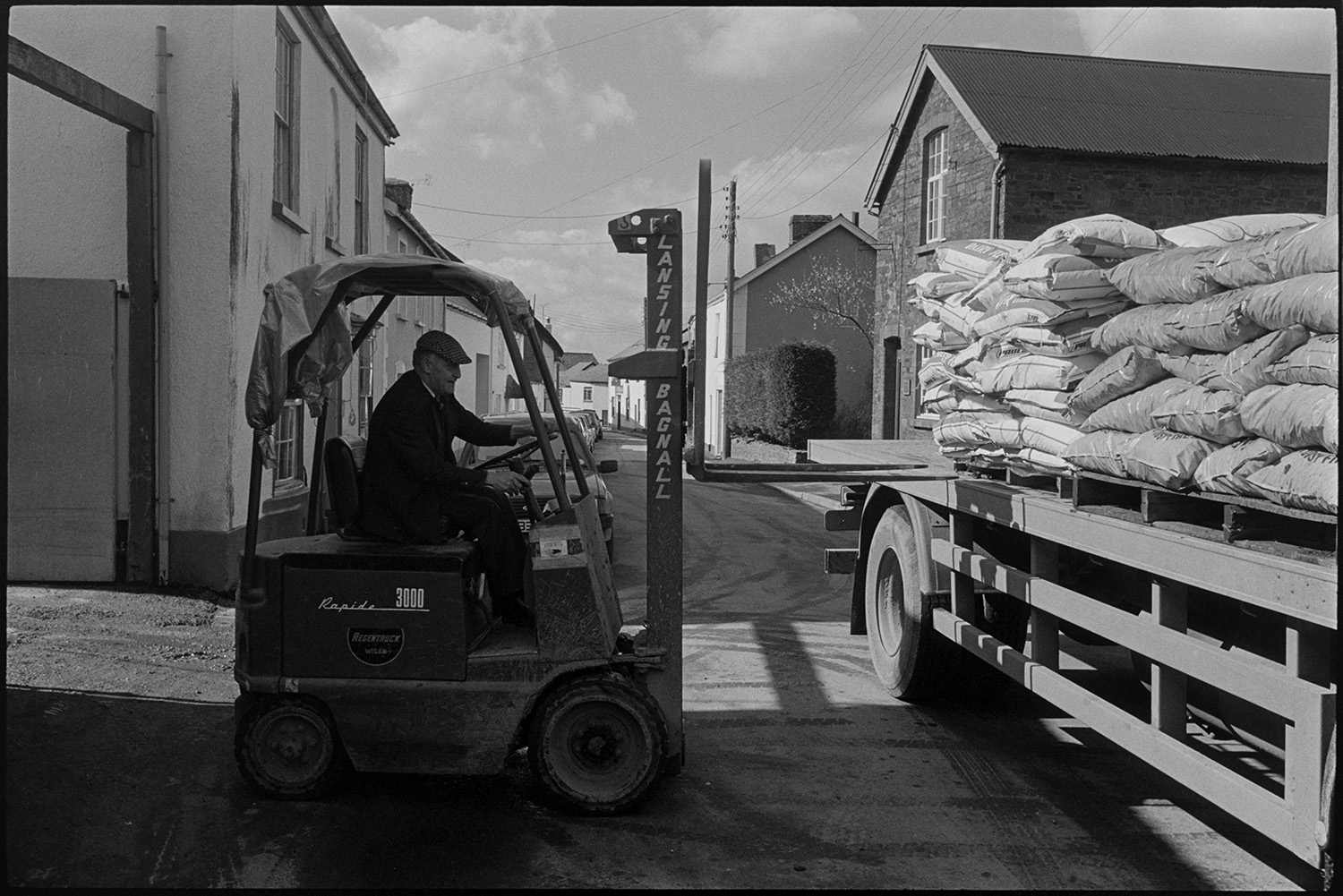 Men unloading fertiliser with fork lift truck.
[Les Shapland using a fork lift truck to unload bags of fertiliser from a lorry parked on East Street, Chulmleigh. There are parked cars and town houses in the background.]