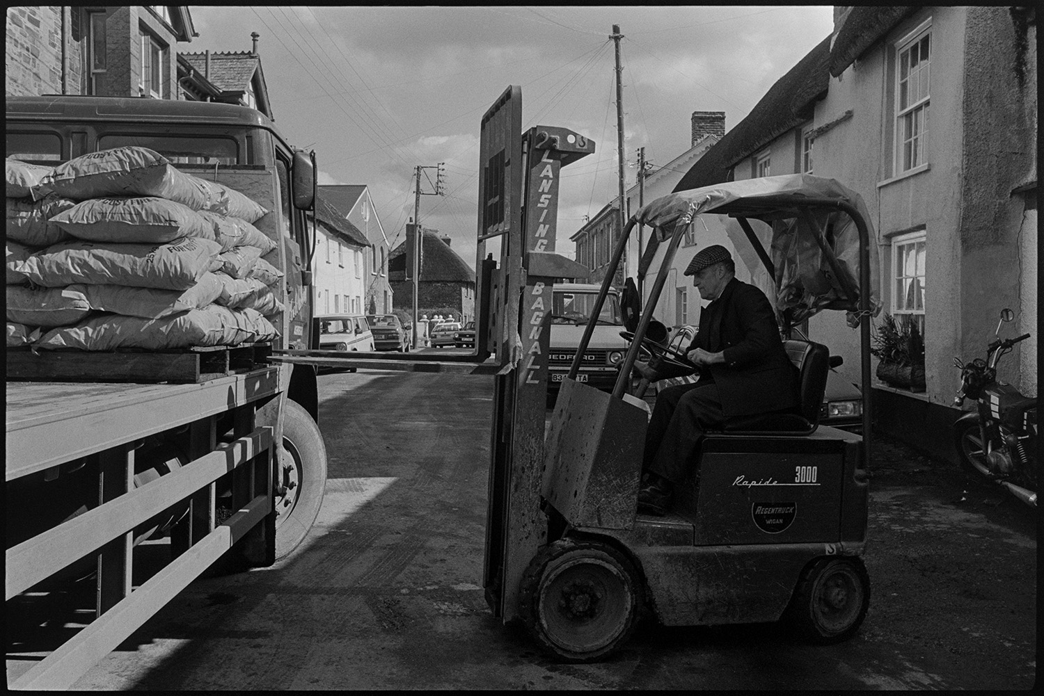 Men unloading fertiliser with fork lift truck.
[Les Shapland using a fork lift truck to unload a pallet with bags of fuel from a lorry parked on East Street, Chulmleigh. There are parked cars and town houses in the background.]