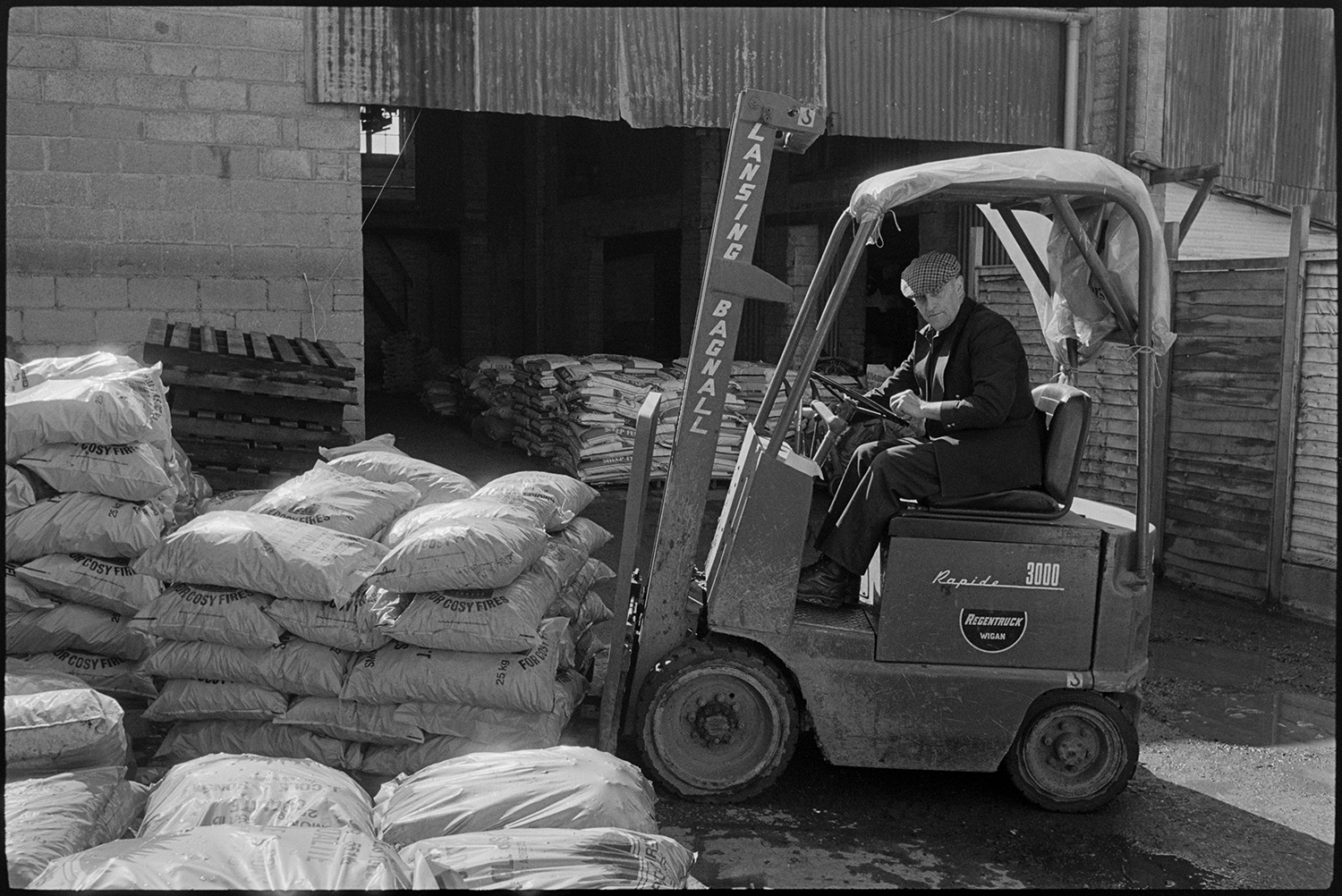 Men unloading fertiliser with fork lift truck.
[Les Shapland is using a fork lift truck to move bags of fuel in a yard at East Street, Chulmleigh. There are more plastic bags stacked on pallets in the corrugated iron shed in the background.]