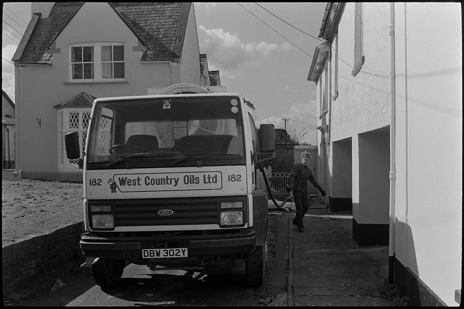 Tanker delivering oil to house.
[A man taking a hose from a parked tanker to deliver oil to a house in New Street, Chulmleigh. The tanker belongs to West Country Oils Ltd. There are other houses in the background.]